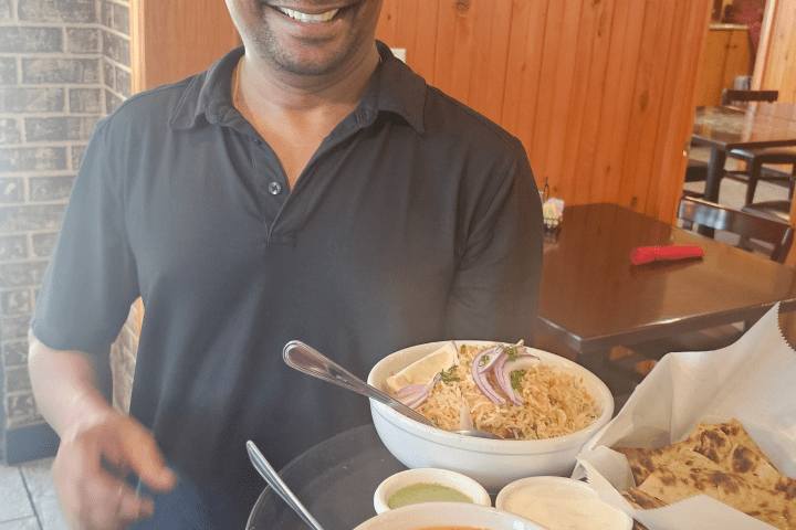 Smiling man holding a tray with curry, rice, naan, and sauces in a restaurant.