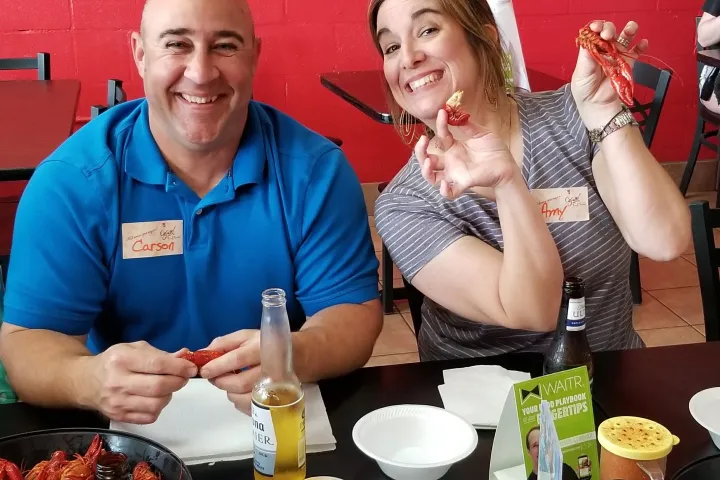 Two people smiling while eating crawfish at a table with drinks and napkins.