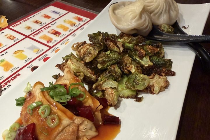 Dumplings, roasted Brussels sprouts, and meat with sauce on a rectangular plate.