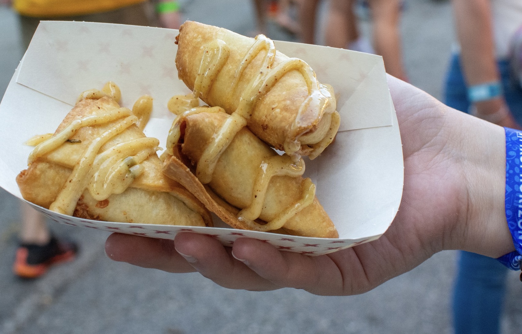 Hand holding tray with three pastries drizzled in sauce outdoors.